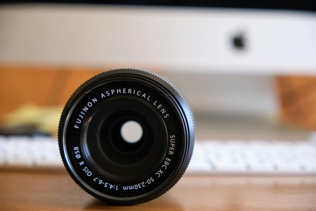 Close-up of a Fujinon aspherical lens with a blurred computer in the background, on a wooden desk.